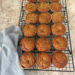 Fresh milled flour banana bread muffins cooling on a baking rack with a blue kitchen towel underneath, golden brown with domed tops and a soft, tender crumb, made with ripe bananas for a wholesome homemade look.