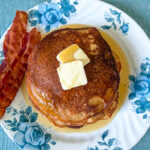 Landscape view of a stack of einkorn flour pancakes on a blue floral plate, served with butter, maple syrup, and two strips of bacon