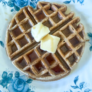 Einkorn waffles with two pats of butter and maple syrup being poured onto it from the top.