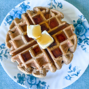 Einkorn waffles with two pats of butter and a stream of maple syrup being poured. Waffles are on a white plate with blue florals on a blue placemat.