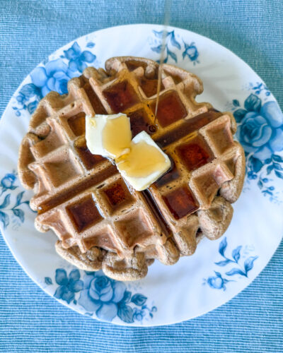 Einkorn waffles with two pats of butter and a stream of maple syrup being poured. Waffles are on a white plate with blue florals on a blue placemat.