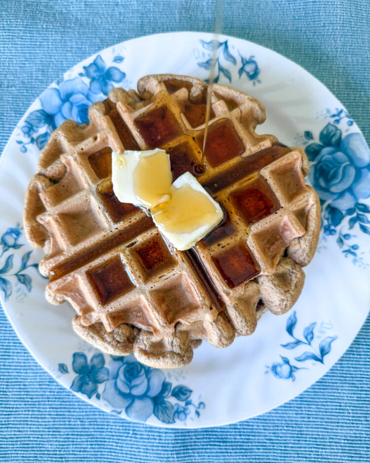 Einkorn waffles with two pats of butter and a stream of maple syrup being poured. Waffles are on a white plate with blue florals on a blue placemat.