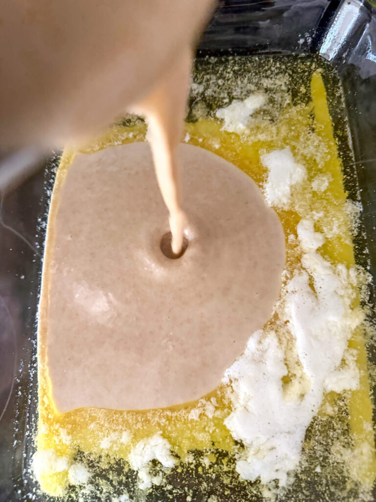 peach cobbler with fresh milled flour batter being poured into a glass baking dish containing melted butter