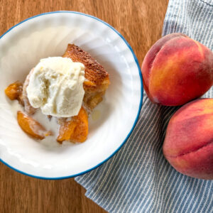 a serving of peach cobbler recipe with fresh milled flour in a blue and white bowl with ice cream on top next to a blue and white striped napkin with two peaches on it