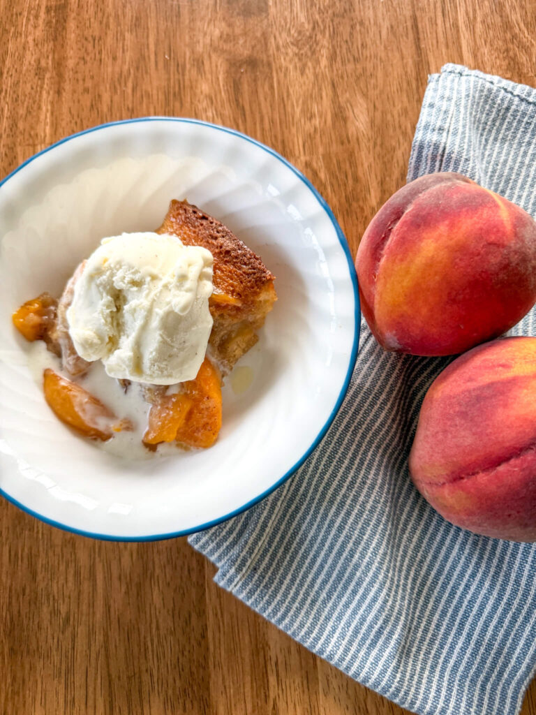 a serving of peach cobbler recipe with fresh milled flour in a blue and white bowl with ice cream on top next to a blue and white striped napkin with two peaches on it