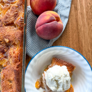 freshly baked peach cobbler recipe with fresh milled flour on wooden table next to two peaches on blue and white striped kitchen napkin and a serving of cobbler in a bowl with vanilla ice cream on top next to it