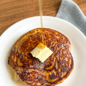 Pumpkin pancake on white plate with pat of butter and maple syrup being poured on top of it.