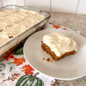 A square of fresh milled flour pumpkin cake on a white plate on top of a pumpkin themed kitchen towel next to a glass baking dish of cake.