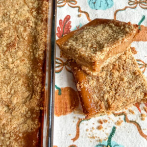 Glass dish of pumpkin pie bars on top of a pumpkin themed kitchen towel next to two pumpkin pie bars stacked on top of eachother.